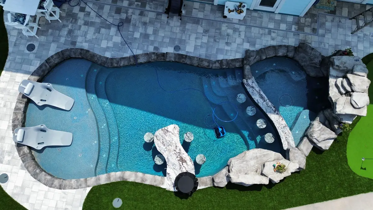 High-angle drone view of a custom backyard lagoon swimming pool with in-water lounge chairs, swim-up bar stools, and waterfall features